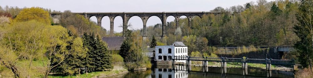 Steinviadukt über einem Flusstal mit einem kleinen weißen Gebäude am Ufer, umgeben von Bäumen und bewaldeten Hügeln.