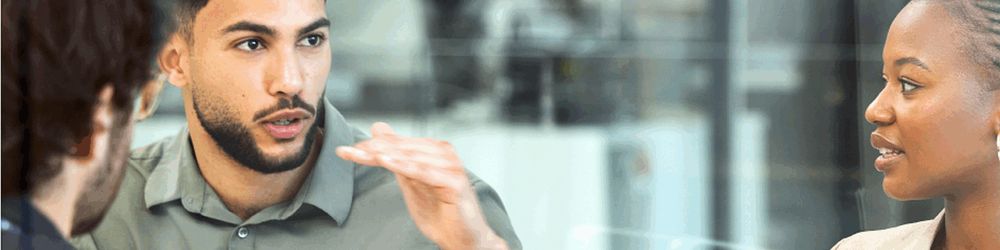 „Drei Personen in einem Gespräch in einem modernen Büro, eine Person spricht und gestikuliert, die anderen hören aufmerksam zu.“