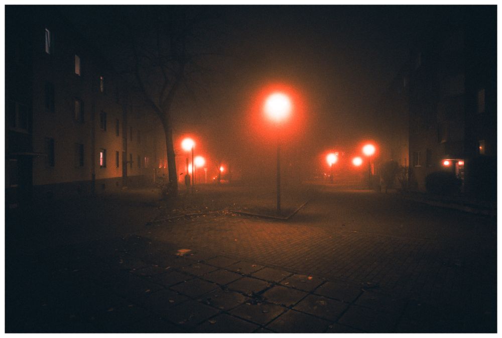photograph of a nighttime street. it's very foggy, and the streetlamps leading down the road all have red-glowing halos. most of the windows in the buildings lining the street are dark, but some emit an eerie glow. the image is noisy and dark where it isn't red-tinted. a few barren trees add to the desolate, creepy atmosphere.