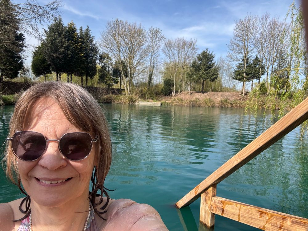 Selfie of Yvonne, a white woman with light brown hair and sunglasses with a view of a blue lake in the background.