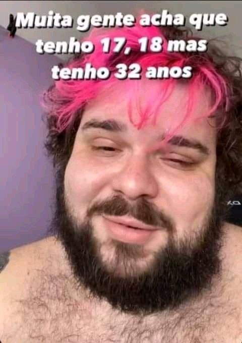 Uma foto de uma pessoa com a franja rosa e barba cheia sem camisa e o texto "muita gente acha que tenho 17, 18 mas tenho 32 anos