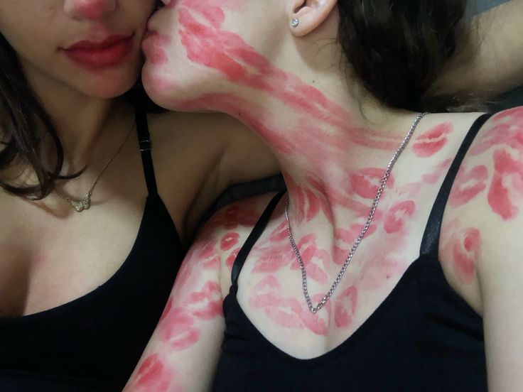 a picture of two girls with black tank tops are seen sharing an intimate moment together. the girl on the left is slightly smiling and the girl on the right is completely covered in lipstick marks as she's leaned in to kiss the other girl