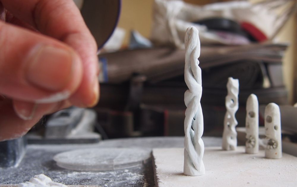 A small, white abstract sculpture that is carved out of a stick of blackboardchalk is just to the right of centre of frame. Slightly out of focus in the background on the right are a few more pieces in the same material. At left, also out of focus, a hand looms into frame, fingers poised as if to pick up or perhaps after putting down the sculpture.