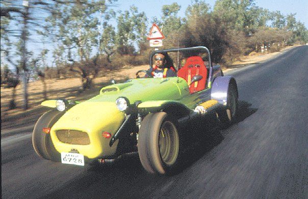 A two-seater roadster coasting down an open, rural road. The vehicle is light yellow and light green. The driver wears dark glasses and seems to have his hair flying in the wind.