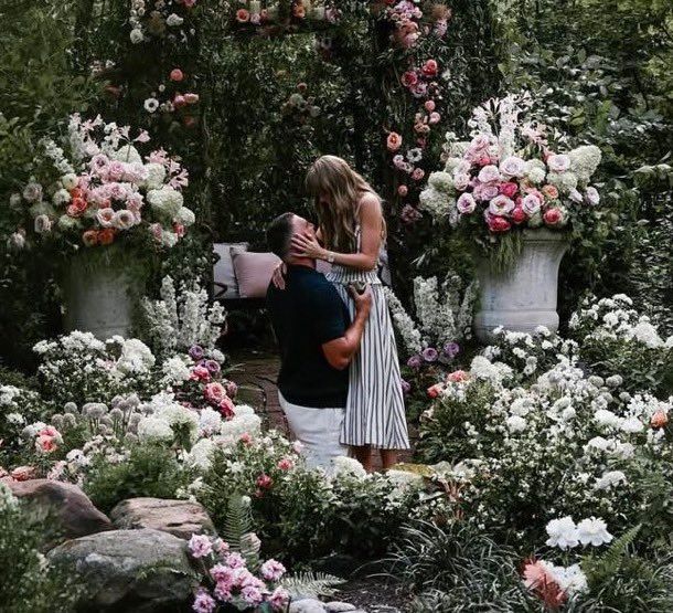 Travis kelce proposing to Taylor Swift. theres flowers and greenery all around them. Taylor is wearing a halter vertical striped dress and Travis is wearing a black top and white pants