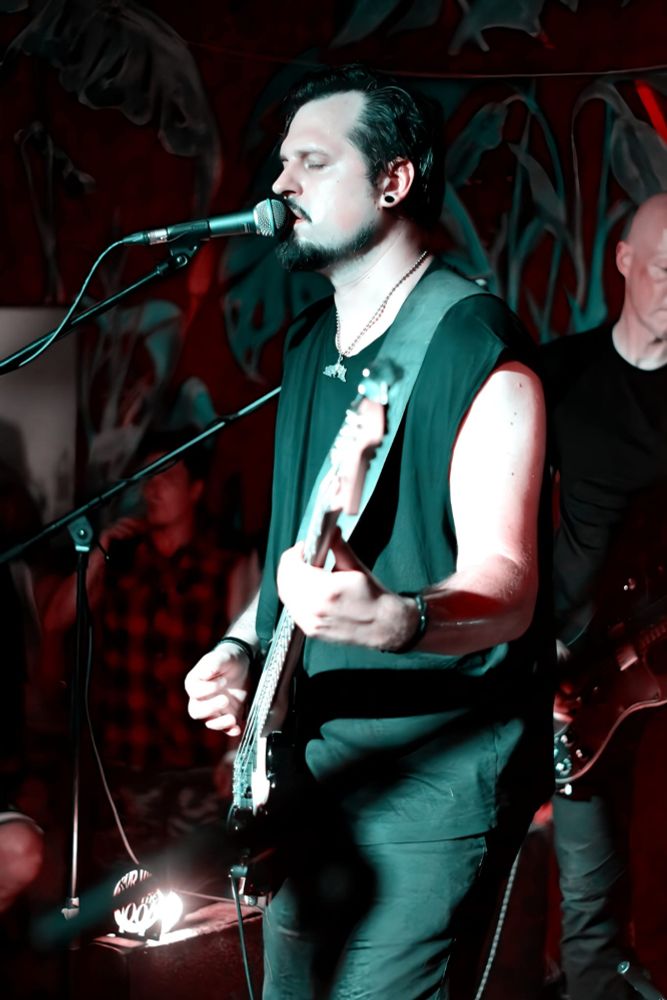 Musiker mit schwarzem ärmellosem Shirt spielt E-Bass und singt mit geschlossenen Augen in Mikrofon auf Bühne. Karol