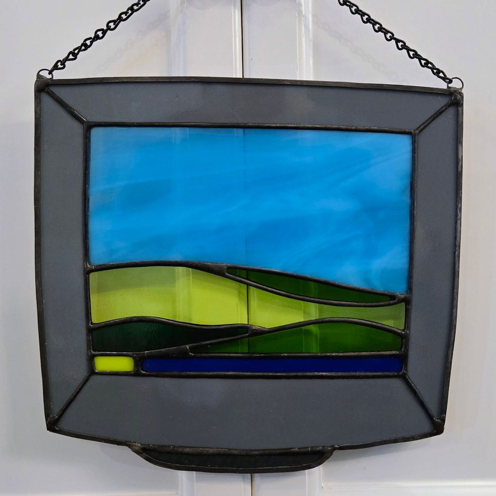 A stained glass suncatcher of an old computer monitor with the windows XP desktop (a lightly cloudy blue sky and rolling green hills) on it