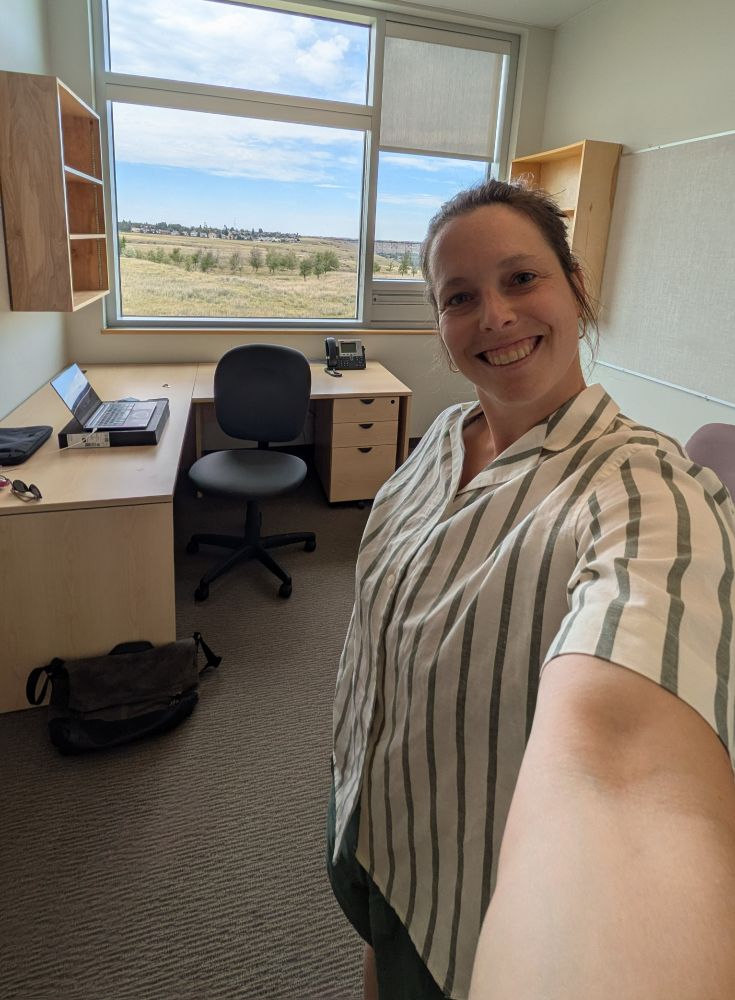 A smiling woman wearing a white and green shirt stands in an empty office with a window overlooking a field and a blue sky, with a single laptop on top of a desk and empty bookshelves. 
