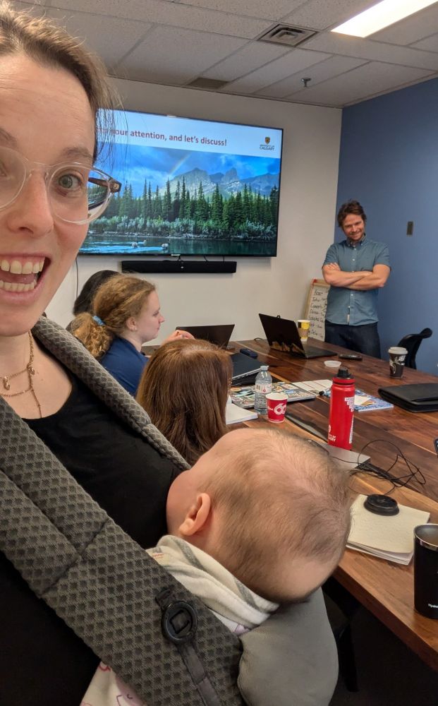 Women carrying a baby in a carrier in a small meeting room with other scientists, with a presenter in the background. 