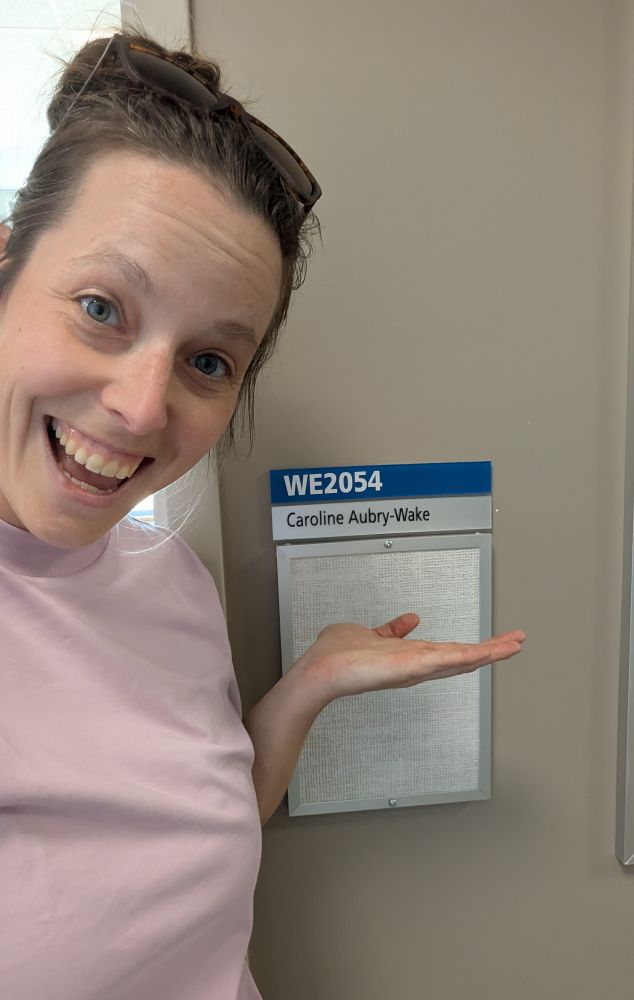 Smiling women in a pink tshirt showcasing her office door with her name on it, being extremely happy about it.
