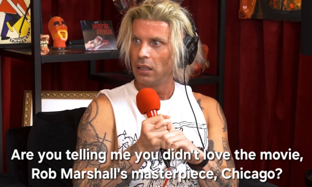 Katya Zamo wears a messy blonde wig, headphones, and a sleeveless white shirt, while holding a red microphone. Her facial expression looks in disbelief. White subtitles on the image read: “Are you telling me you didn’t love the movie, Rob Marshall’s masterpiece, Chicago?”