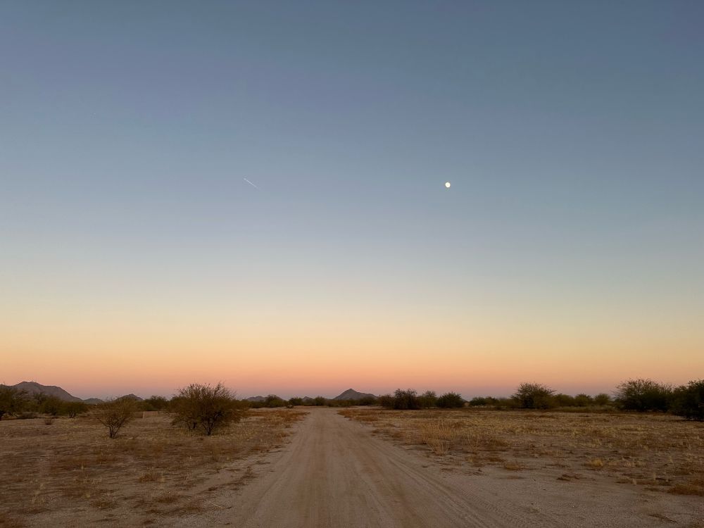 along the bottom is a yellowy brown feild of dirt and dry grass with an occasional green/brown bush, in the very distance there are mountains and in the middle is a dirt road,, along the horizon a rainbow of colors and starting at the horizon its going from purple to redish orange to yellow, slight green and then up into the light blue sky,, in the sky on the left middle side at the same level of the moon is a very very faint airplane leaving a streak and the right middle side is the moon glowing as a small white dot