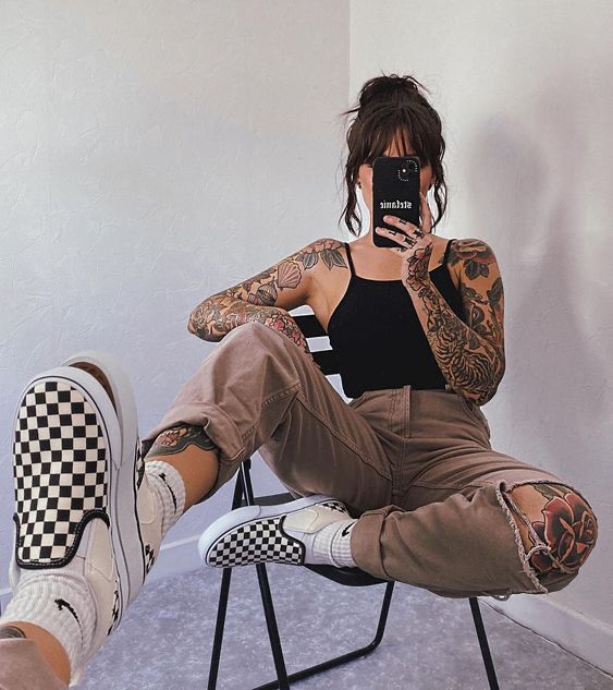 Heavily tattooed woman wearing black tank top along with brown pants with holes and checkered black and white vans.