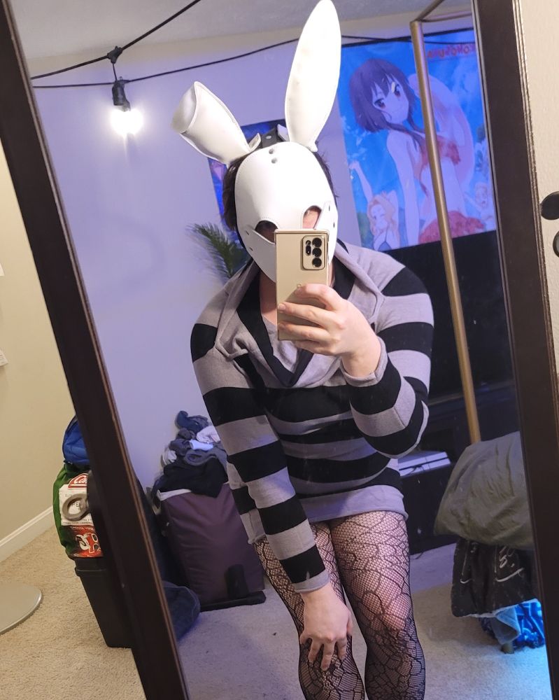 Femboy wearing a white leather bunny hood, black and grey striped dress, and spiderweb fishnets leaning forward taking a cute selfie. 
