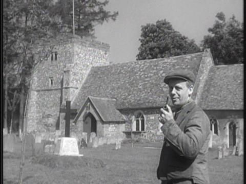A man holds a pipe while looking at the camera, in front of an English village church 