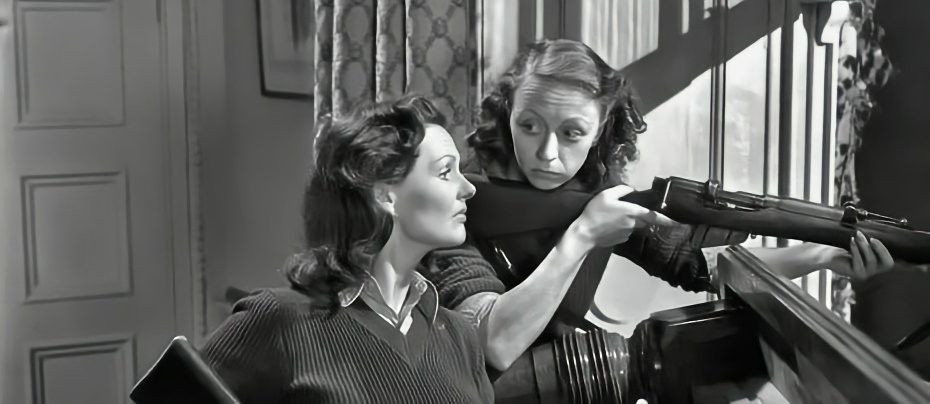 A young woman holding a rifle pointing out of a window looks away from its sight to an older woman by her side, whose gaze is set on the rifle's target. 