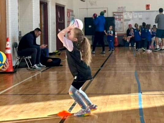 A 5-year-old girl with long hair in a ponytail sets up her shot at basketball practice. She is dressed in a black basketball jersey and wears light blue coloured snug leggings with pastel rainbow stars and hearts. Her shoes have a rainbow gradient on the sole and unicorns on the side. Her arms are bent above her head, and she's basically willing her pink, purple, and turquoise ball to make its way to the basket, despite it bring about 7 feet over her head. (Spoiler: the ball is going to bounce off the rim and she will be excited that she hit it. Great attitude, kid!)