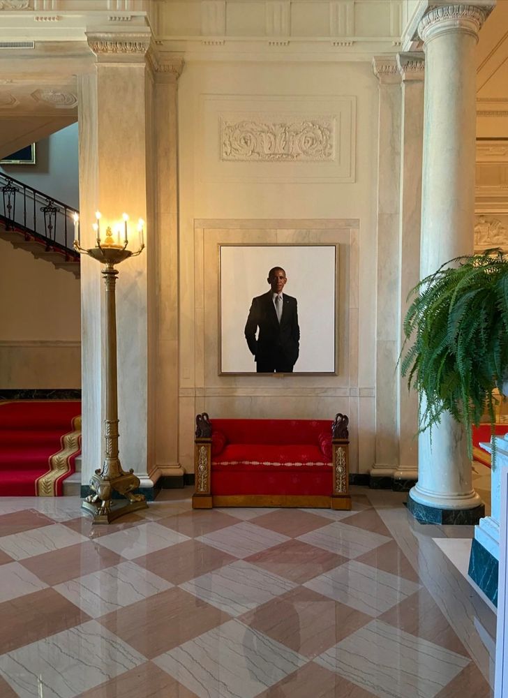 Image of President Obama's official portrait hanging in the White House, above a red upholstered antique bench. The portait's reflection can be seen on the marble floor which is a red and white diamond checkerboard design. To the left pf the portait and bench are a three footed antique brass lamp next to the entrance to a red carpeted staircase. To the right is a large marble column, which along with a marble demi-wall topped with a marble planter containing a lusciously full and vibrant green fern, demarcates the entrance into an adjoining space.