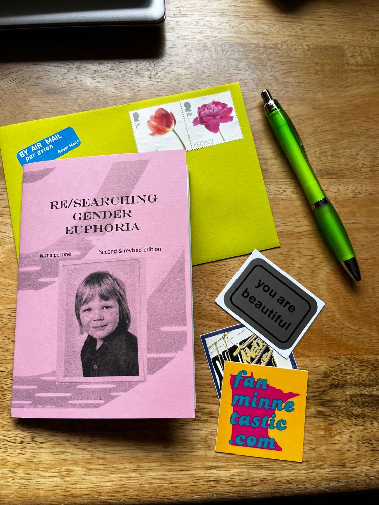 A pink zine called Re/searching gender euphoria, a green envelope with flower postage stamps, a green pen, three stickers. 