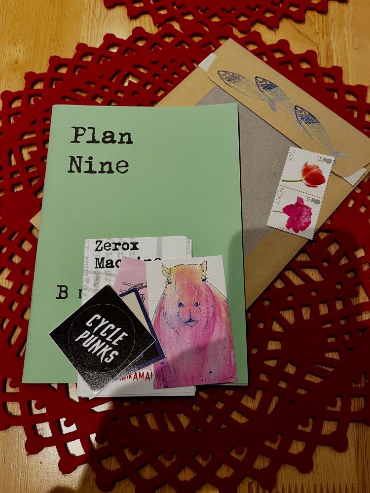 A pale green A5 zine with the title Plan nine with assorted stickers including a handdrawn capybara. Flower-themed UK postage stamps. All on a red felt place mat on a wooden table. 
