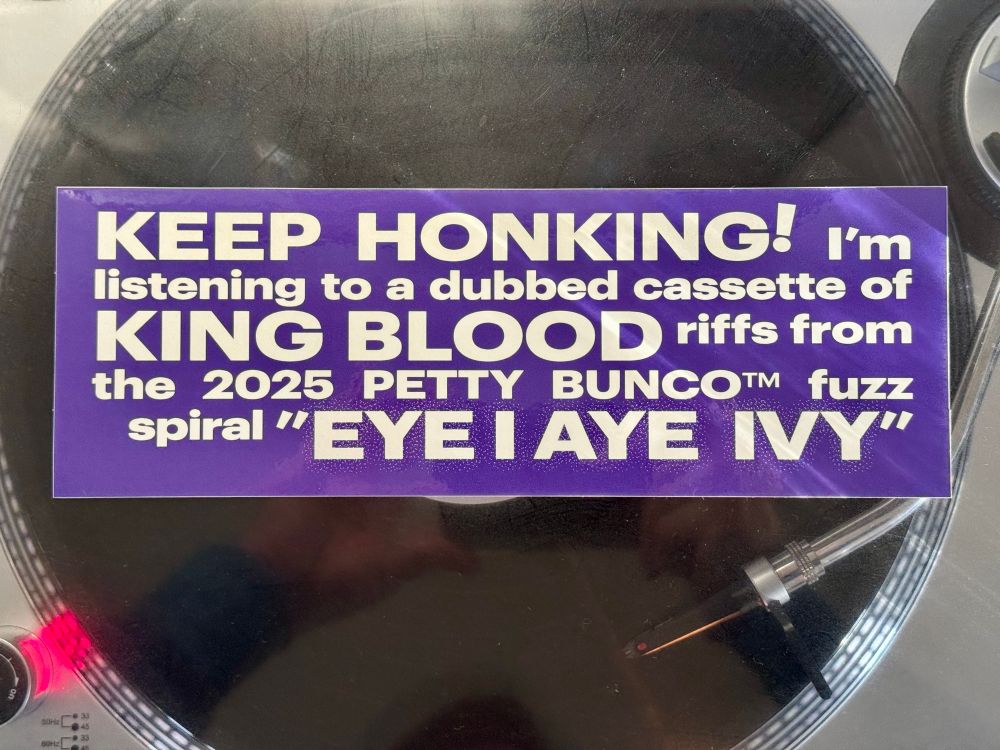 Bumper sticker that reads “KEEP HONKING! I'm listening to a dubbed cassette of KING BLOOD riffs from the 2025 PETTY BUNCO™ fuzz spiral "EYE I AYE IVY"” sitting atop the dust cover of a record player while a record plays on the turntable it’s covering.