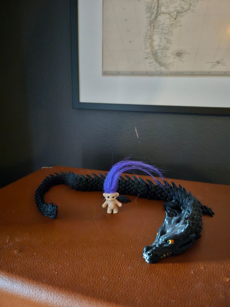um trollzinho de cabelo roxo em pé em cima de uma toca-discos portátil fechado e um dragao preto articulado ao lado