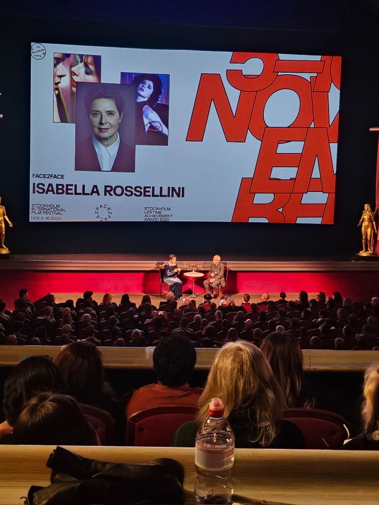 sala de cinema exibindo um painel na tela com o nome da atriz e embaixo, em carne e osso, ela mesma está conversando, sentada em uma cadeira