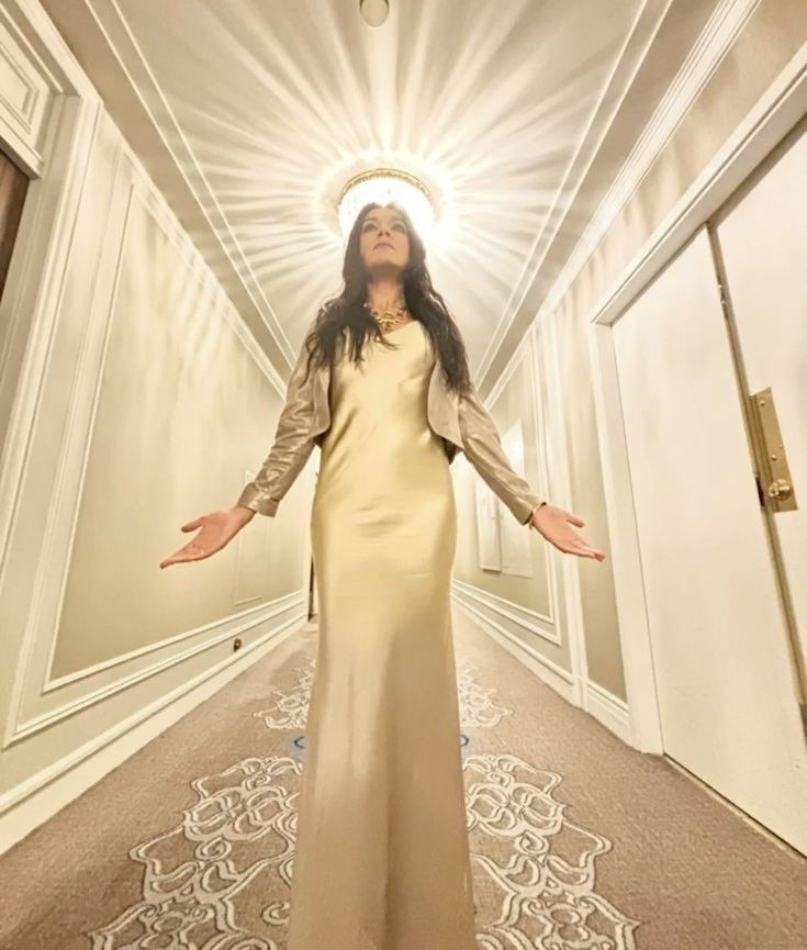 woman in a long white dress in a hotel corridor. she has her palms up as if praying and above her the light fixture looks like haloed sun. she has long dark hair and wears some jewelry around her neck. it’s the actress simone kessell