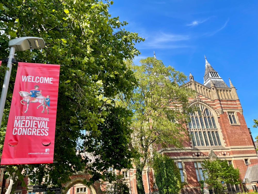 A photo of a red sign that says ‘Welcome Leeds International Medieval Congress’ with The Great Hall in the background. It is a sunny day with a nearly clear sky. 