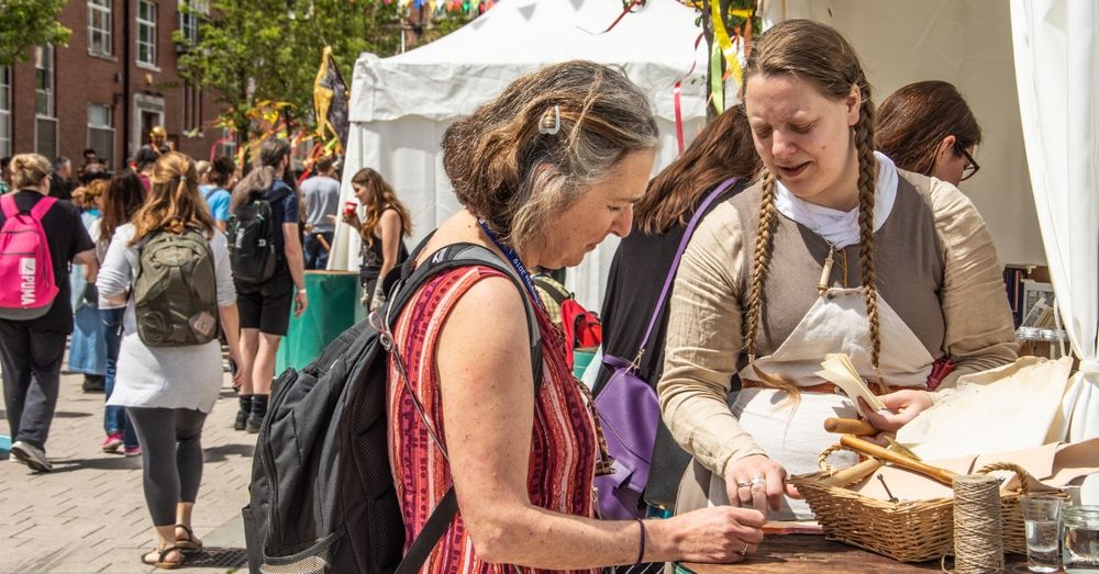 Two people interacting at a market booth at the International Medieval Congress with other visitors and festive decorations in the background.