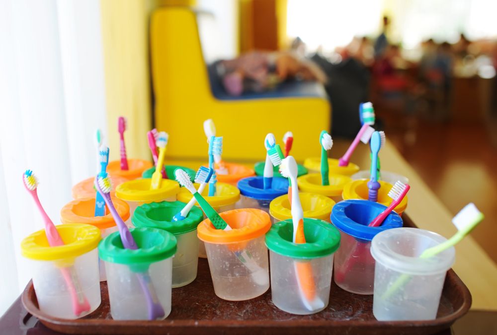 A number of colourful children's toothbrushes in cups on a tray. The background looks like a nursery or a school.
