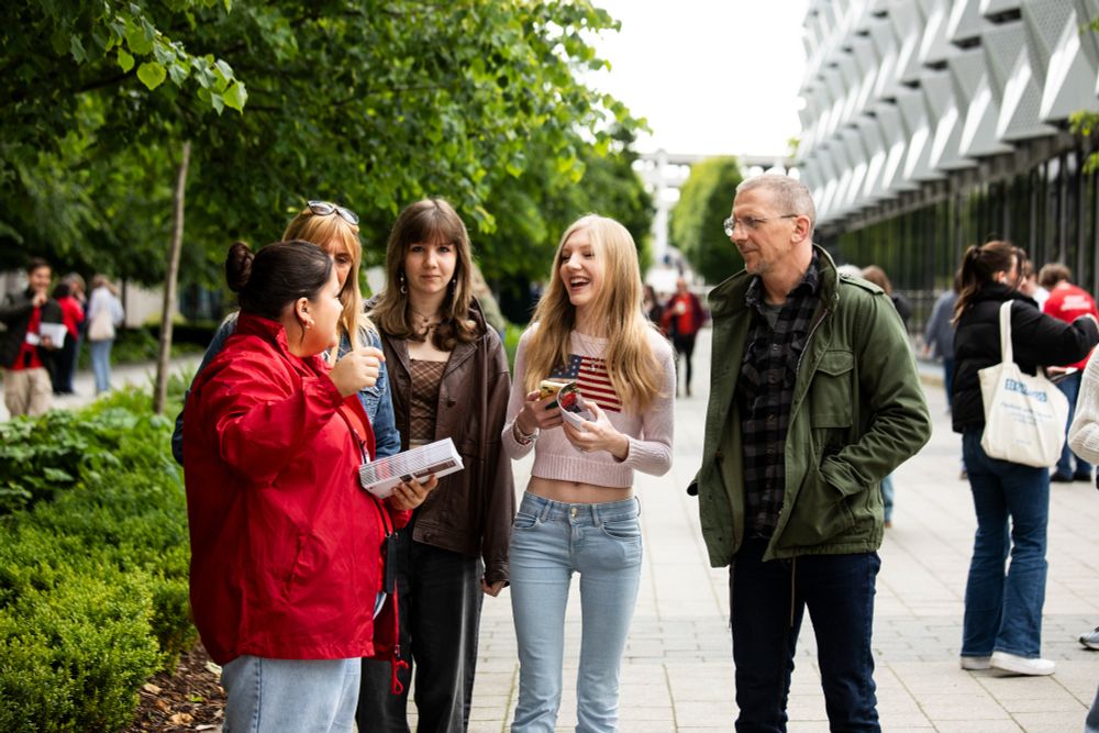 A student ambassador engaging in conversation with four people on campus. The student ambassador holds leaflets and is gesturing with their hands. Trees and the university's multi-storey car park can be seen in the background.