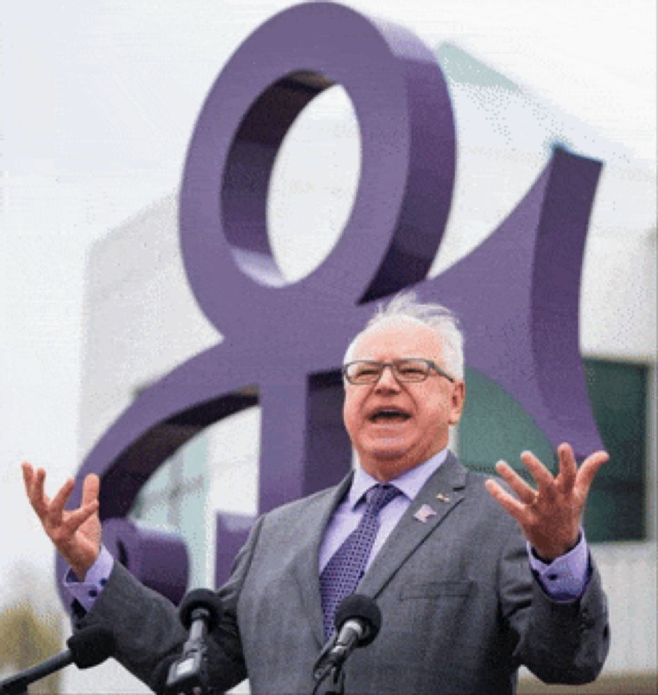 a man in a suit and tie stands in front of two microphones in front of a large purple symbol