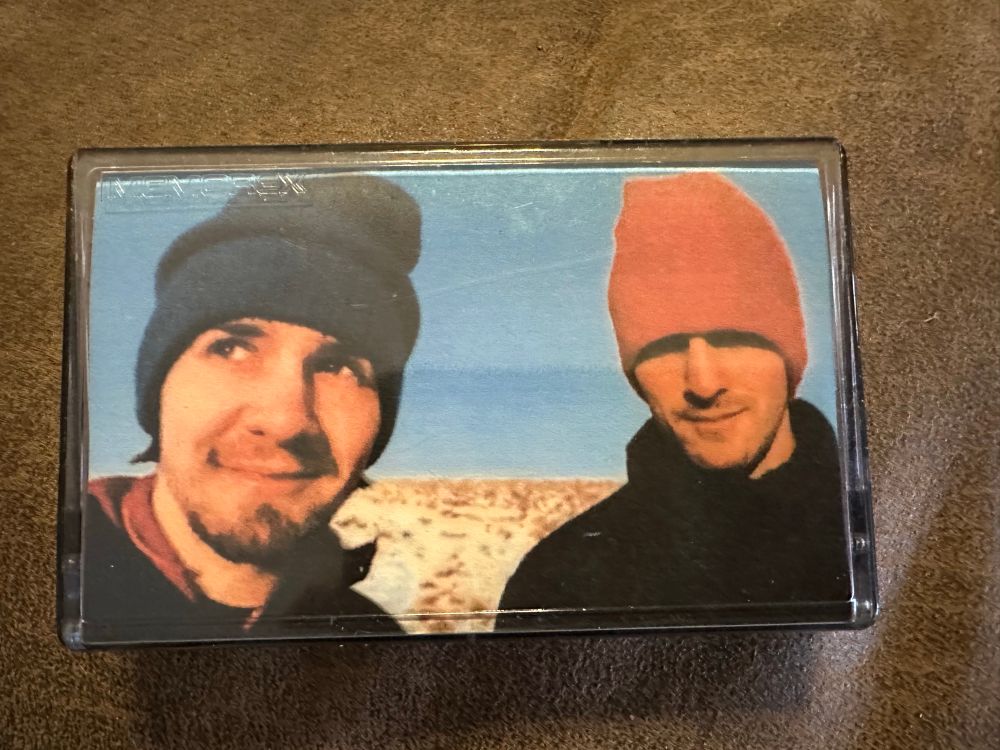 the front of the cassette. an image of Mike Sandison (left) and Marcus Eoin (right) wearing woolen hats (Mike's is blue, Marcus's is red) and black jackets in front of what looks like a barren field covered with a thin layer of snow, or maybe just a light colored grass, under a clear blue sky. they're both smiling slightly, with Mike looking off to the left and Marcus looking directly at the camera.