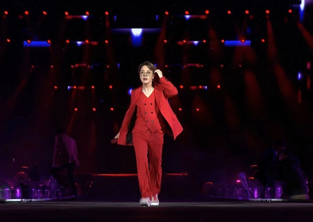 Jhope from BTS walking down a stage wearing a fully red suit with a red vest. The stage lighting behind him is red and blue. He is holding his left hand up towards his ear sweeping his hair behind it
