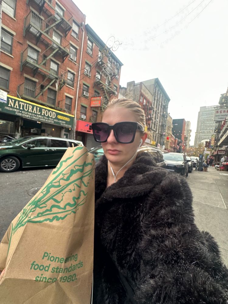 a white woman wearing wired headphones and a brown fur coat carrying a whole foods bag