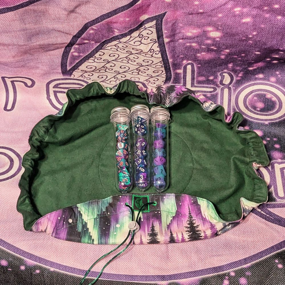 an open drawstring bag made of purple and green fabric with an aurora borealis above a forest print on the outside and a dark green fabric on the inside. three different sets of purple and blue or purple and aqua dice are set in the middle of the bag inside clear plastic tubes.
