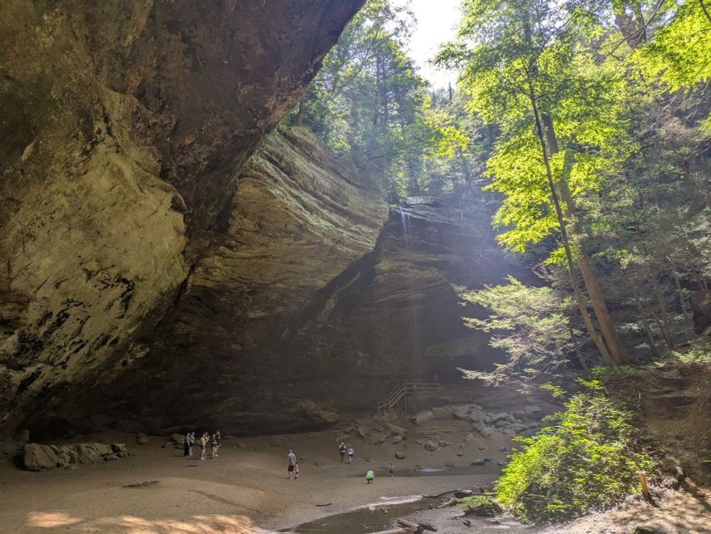 A broad cave with a small waterfall cascading from high above. People stand and admire the scenery. Photo taken at Hocking Hills State Park in Ohio.