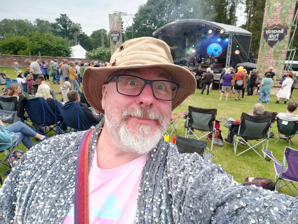 Selfie of a badly shaved potato of a man wearing a brown hat, pink t-shirt, and ridiculously sparkly silver jacket. 
In the background a stage with a band playing on it.