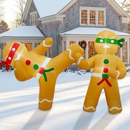 Two gingerbread men with belts and eyemasks like ninjas, one red, one green. They're standing on smooth flat snow in front of a two-story grey house with snow on the roofs. The red ninja is standing on its right leg while kicking green ninja with its left. Green ninja is blocking the kick with its right hand. Red ninja has a HA! expression and green ninja has a ARGH! expression