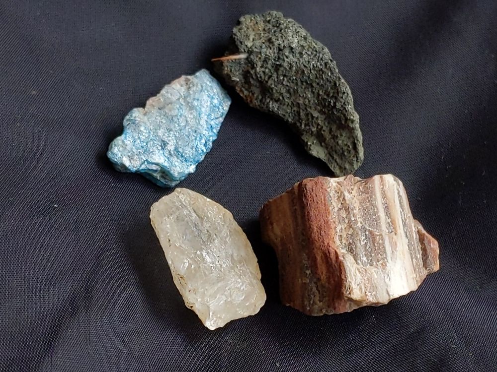 Four rocks on a black fabric with natural light. Descriptions: shiny blue-green, rough charcoal grey with hint of olive green and a small smooth brown bit attached, a white to translucent shiny quartz, and a multi-layered possible sandstone of rust brown to brown to white.