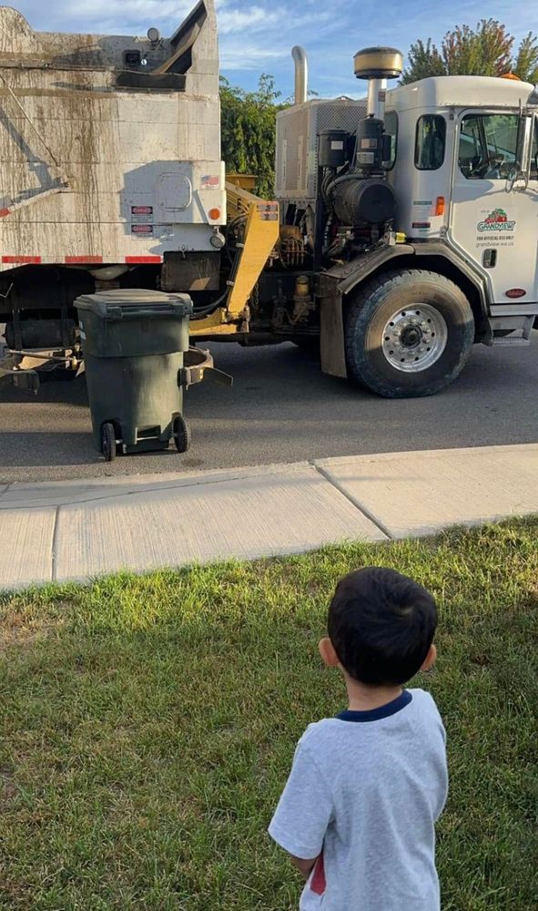 Small child in the foreground standing on their front yard grass watches avidly as trash truck robot arm picks up the trash bin at the curb.