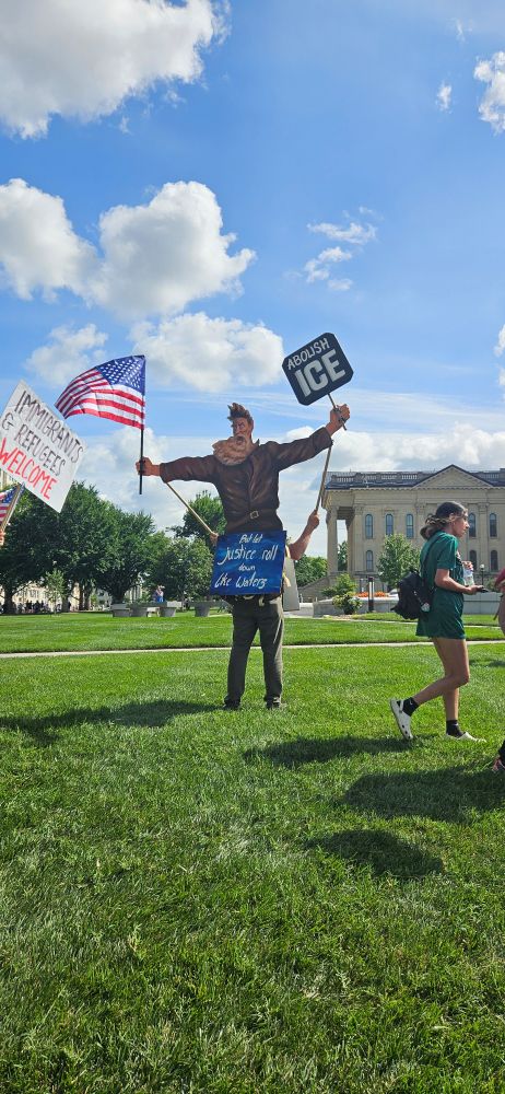 Lifesize cut out of John Brown on the back of a protester. John Brown is holding an American flag in one hand and a sign that says, "abolish ice," in the other hand. At John Browns waist is a sign that says, "but let justice roll down like waters."