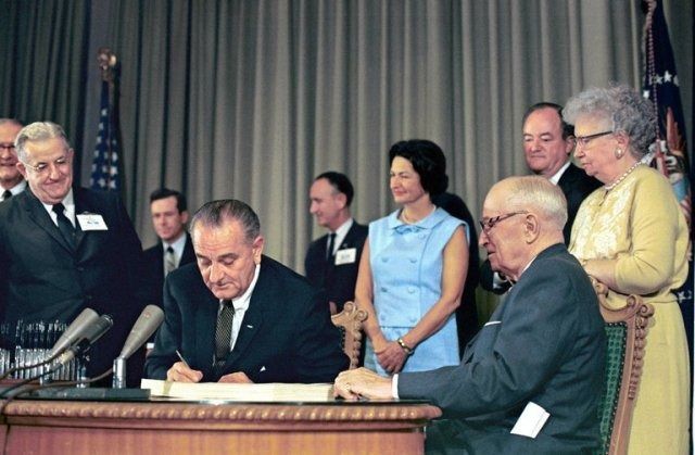 Lyndon Johnson signing the law that created Medicare and Medicaid. His wife looks on in the background, near Vice President Humphrey. In the foreground on the right are Harry and Bess Truman.
