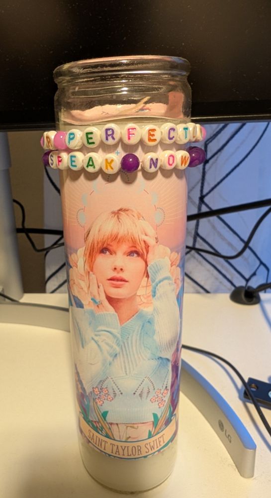 Catholic prayer candle with a Saint Taylor Swift label. It's also adorned with my friendship bracelets "Mr Perfectly Fine" and "Speak Now". It's on a desk in front of a monitor, some cords, and a curtain