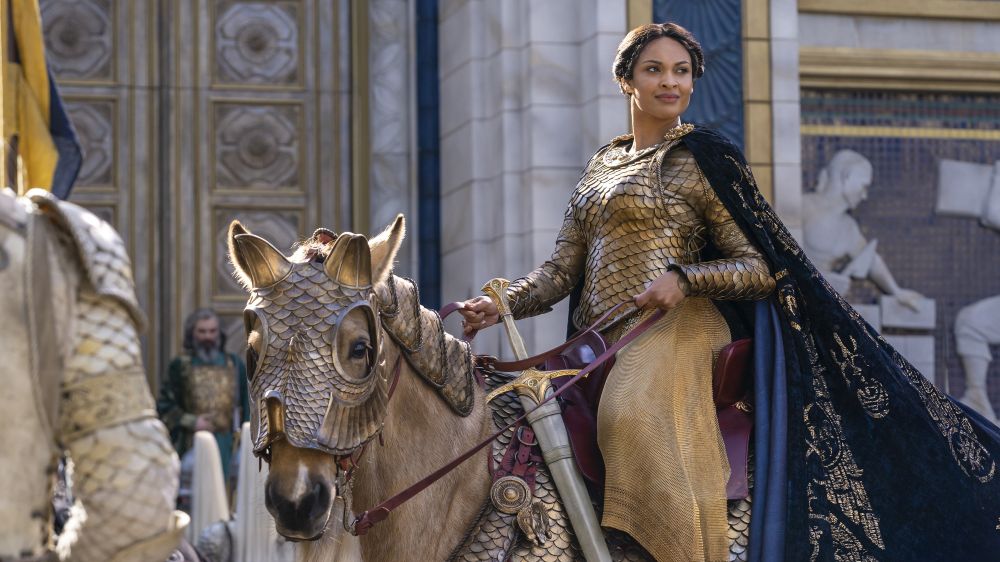 Míriel in The Rings of Power season one on her horse. She is wearing a long, stunning cape with golden details and a breast plate/hauberk resembling golden fish scale. 