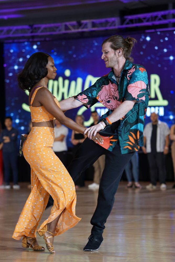 Two people are dancing together on a wooden floor. The person on the left is wearing a yellow, floral-patterned, sleeveless top and matching pants, along with tan dance shoes. The person on the right is wearing a colorful shirt with a floral and abstract design, black pants, and black shoes. The background features a large screen with a blue and purple starry design and partially visible text that reads "Swing" and "Open." There are several people standing in the background, observing the dancers.