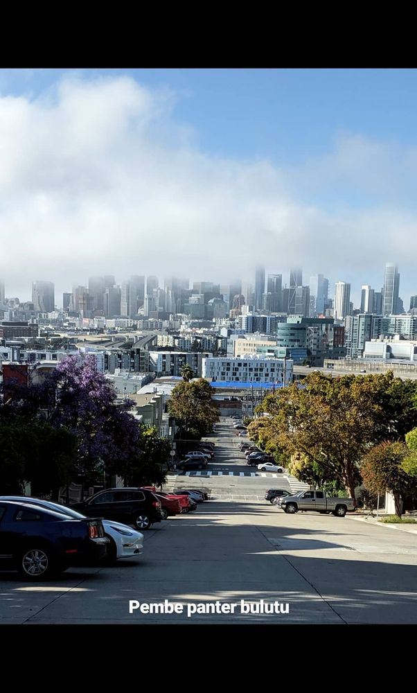 Uzaktan gökdelenlerin olduğu bir şehrin görüntüsü. Yokuş bir cadde fotoğrafın çekildiği yerden şehre doğru uzanıyor. Şehrin, binaların üzerinde bulutlar, sisler. Yüksek binaların tepeleri sisin içinde.