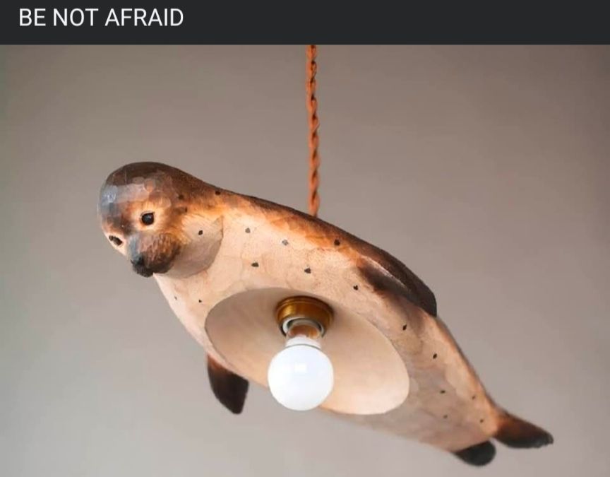 Text above image: BE NOT AFRAID
Image: a hanging wooden light fixture carved and painted brown, black, and beige to resemble a seal, flippers and all. In the middle of its hollowed out belly is a lightbulb. The curious creature dangles from a thin orange cord from the middle of its back.