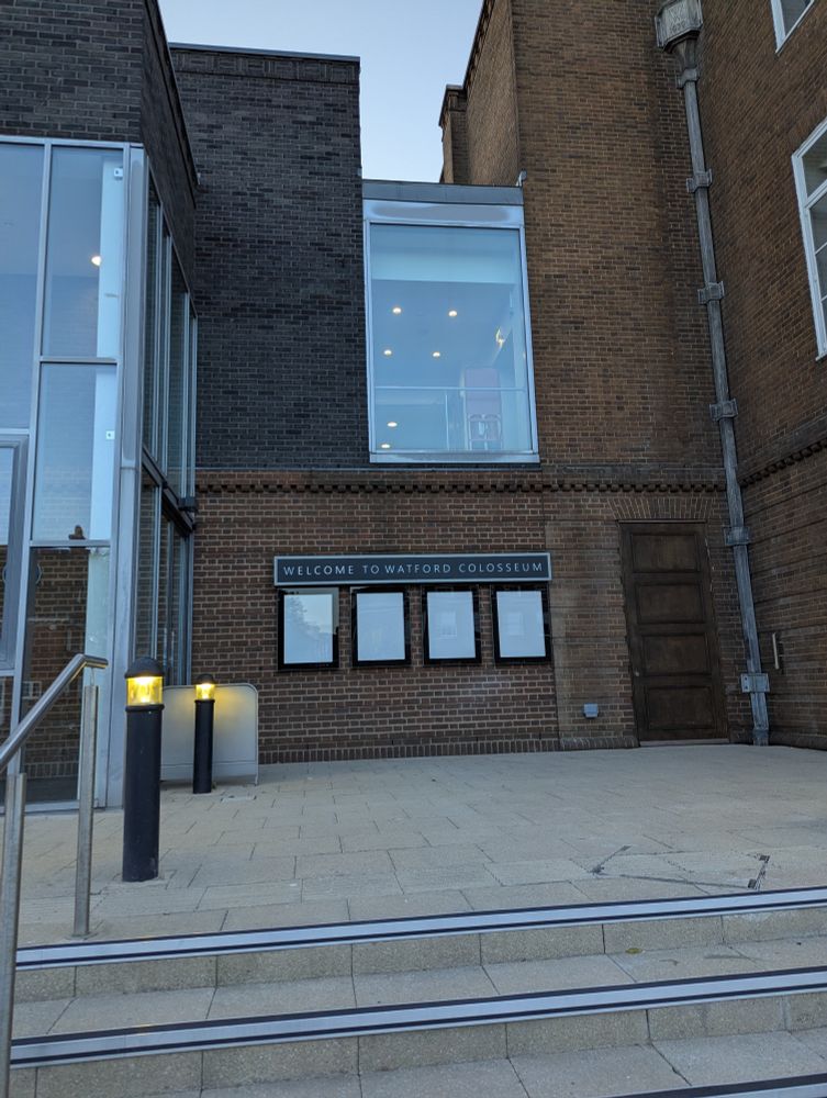 Steps leading up to the entrance nearest the bar/cafe side. A plaque reads 'Welcome to Watford Colosseum'. Under the plaque are 4x empty spaces where billboards for acts would be placed.